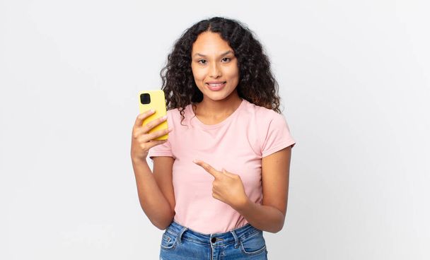 hispanic pretty woman smiling cheerfully, feeling happy and pointing to the side and holding a smartphone - Foto, Imagem