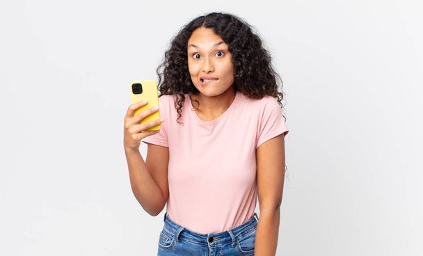 hispanic pretty woman looking puzzled and confused and holding a smartphone - Foto, Bild