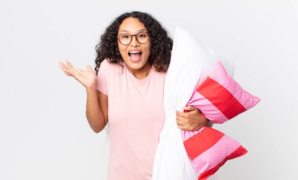 hispanic pretty woman feeling happy and astonished at something unbelievable wearing pajamas and holding a pillow - Foto, immagini