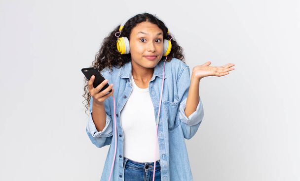 hispanic pretty woman feeling puzzled and confused and doubting and holding a smartphone and headphones - Photo, Image