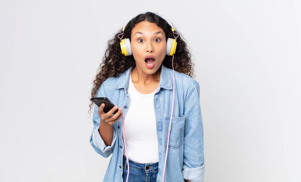 hispanic pretty woman looking very shocked or surprised and holding a smartphone and headphones - Photo, Image