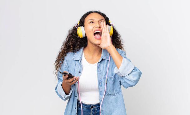 hispanic pretty woman feeling happy,giving a big shout out with hands next to mouth and holding a smartphone and headphones - Photo, Image