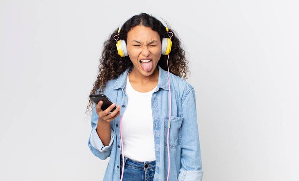 hispanic pretty woman with cheerful and rebellious attitude, joking and sticking tongue out and holding a smartphone and headphones - Photo, Image