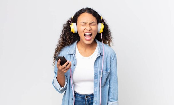 hispanic pretty woman shouting aggressively, looking very angry and holding a smartphone and headphones - Photo, Image