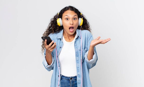 hispanic pretty woman amazed, shocked and astonished with an unbelievable surprise and holding a smartphone and headphones - Zdjęcie, obraz