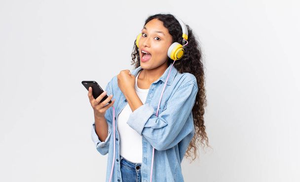 hispanic pretty woman feeling happy and facing a challenge or celebrating and holding a smartphone and headphones - Photo, Image