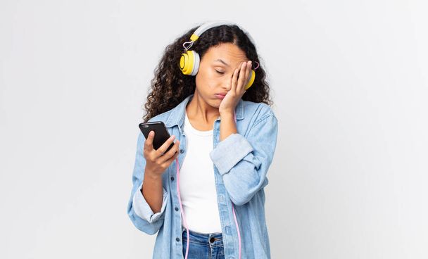hispanic pretty woman feeling bored, frustrated and sleepy after a tiresome and holding a smartphone and headphones - Photo, Image