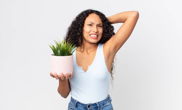 hispanic pretty woman feeling stressed, anxious or scared, with hands on head and holding a houseplant - Fotografie, Obrázek