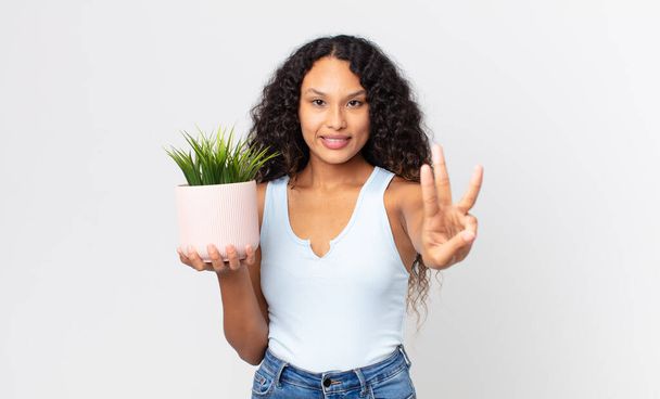 hispanic pretty woman smiling and looking friendly, showing number three and holding a houseplant - Foto, immagini