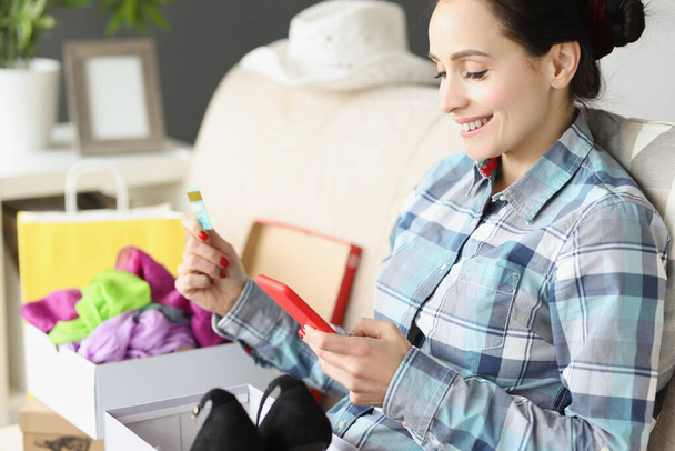 Uśmiechnięta kobieta trzyma kartę bankową i smartfona siedząc na kanapie z pudełkiem buta - Zdjęcie, obraz