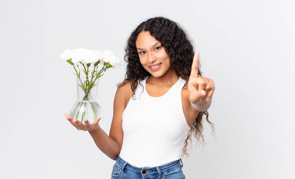 hispanic pretty woman smiling proudly and confidently making number one and holding a flowers jar - Photo, Image