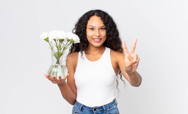 mujer bonita hispana sonriendo y buscando amigable, mostrando el número dos y sosteniendo un frasco de flores - Foto, imagen