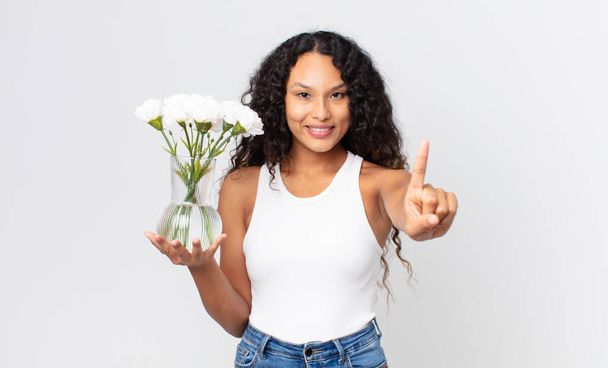 hispanic pretty woman smiling and looking friendly, showing number one and holding a flowers jar - Photo, Image