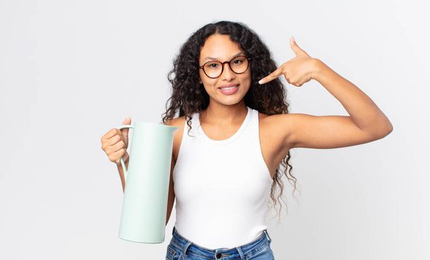 hispanic pretty woman smiling confidently pointing to own broad smile and holding a coffee thermos - Foto, Bild