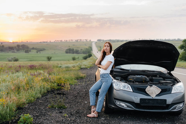 A frustrated young girl stands near a broken-down car in the middle of the highway during sunset. Breakdown and repair of the car. Waiting for help. - Fotografie, Obrázek