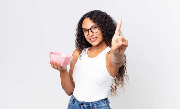 hispanic pretty woman smiling proudly and confidently making number one and holding an empty pot - Foto, Imagem