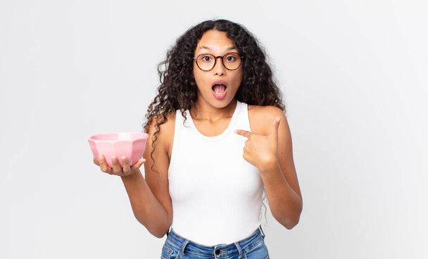 hispanic pretty woman looking shocked and surprised with mouth wide open, pointing to self and holding an empty pot - Foto, Bild