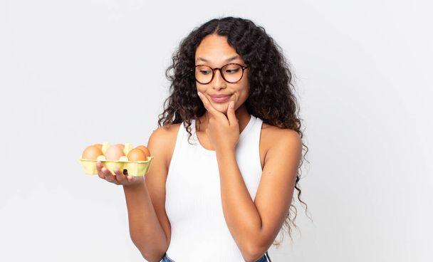 hispanic pretty woman smiling with a happy, confident expression with hand on chin and holding an eggs box - Photo, image