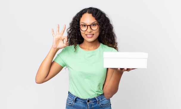 hispanic pretty woman feeling happy, showing approval with okay gesture and holding a white box - Фото, изображение