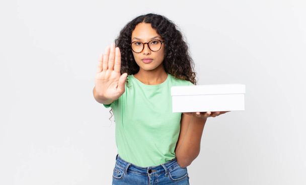 hispanic pretty woman looking serious showing open palm making stop gesture and holding a white box - Foto, Bild