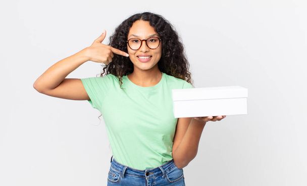 hispanic pretty woman smiling confidently pointing to own broad smile and holding a white box - Foto, afbeelding