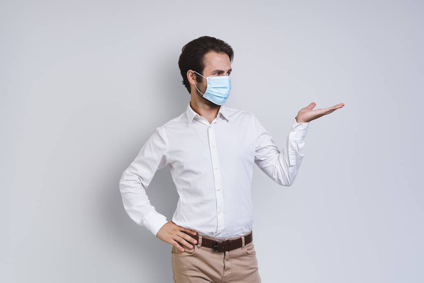Confident young man in white shirt wearing protective face mask and gesturing while standing against gray background - Photo, image