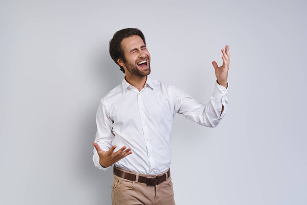 Happy young man in white shirt gesturing and keeping eyes closed while standing against gray background - Фото, изображение