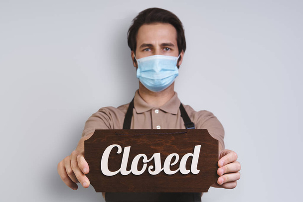 Confident young man in apron wearing protective face mask and holding close sign while standing against gray background - Foto, imagen