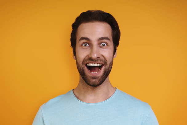 Portrait of excited young man in casual clothing looking at camera and smiling while standing against yellow background - Foto, Imagen