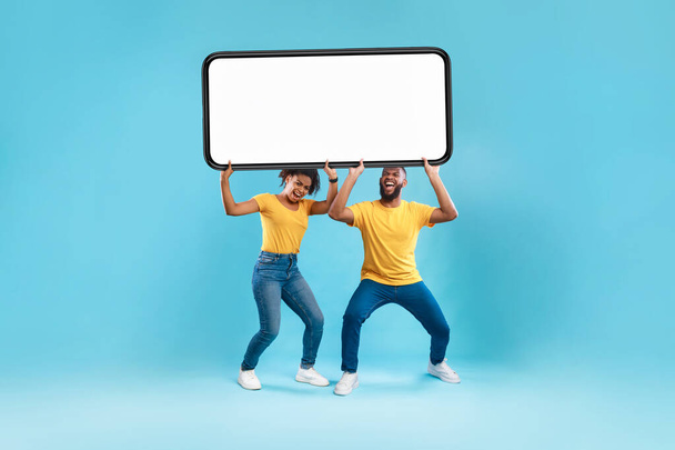 Schwarzes Paar mit riesigem, schwerem Smartphone mit leerem Bildschirm auf blauem Hintergrund. Mockup für Werbung, mobile App oder Website - Foto, Bild