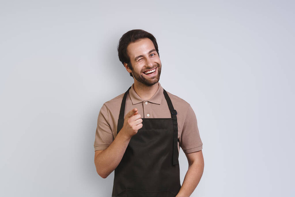 Handsome young man in apron looking at camera and pointing you while standing against gray background - Foto, imagen