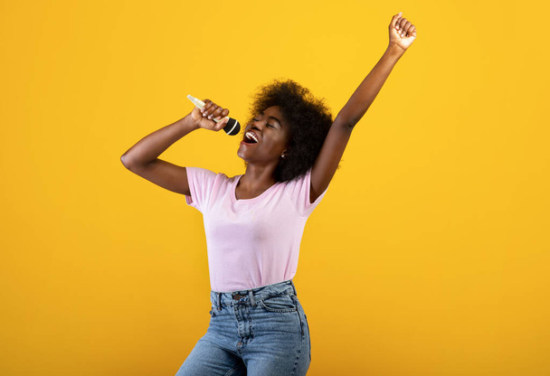 Music lover. Excited black woman singing songs in microphone and dancing, having fun over yellow background - Фото, зображення