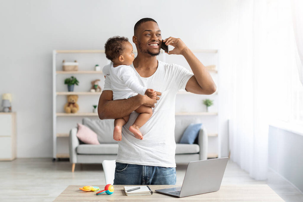 自宅で働く中で携帯電話で話す赤ちゃんを持つ黒人男性 - 写真・画像