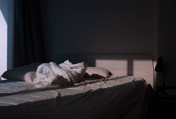 An unmade empty bed in the dramatic dim light coming from a window - Foto, Imagen