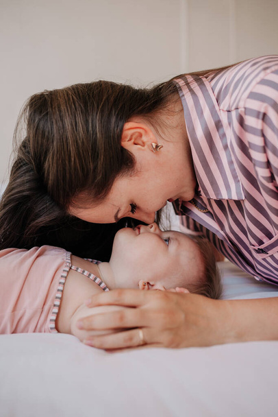 brunette mom in pajamas hugs a baby girl on the bed in the bedroom - Фото, изображение