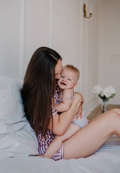 beautiful brunette mom in pajamas hugs her baby daughter on the bed in the room - Photo, Image