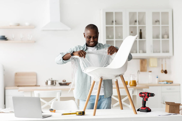 Happy black man receiving new furniture and start unpacking chair on table in kitchen interior, copy space - Photo, Image