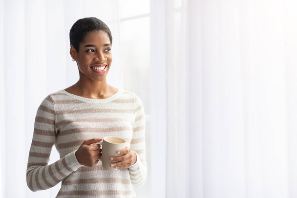 Šťastný úsměv afroameričanka pití kávy, zatímco stojí v blízkosti okna doma - Fotografie, Obrázek
