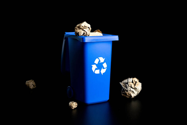 Separacja papieru recyklingu. Niebieski kosz na śmieci do recyklingu papierowych śmieci odizolowanych na czarnym tle. Pojemnik na śmieci do unieszkodliwiania odpadów i ochrony środowiska - Zdjęcie, obraz