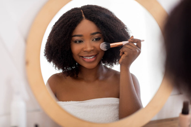 Mooie zwarte dame aanbrengen make-up in de buurt van spiegel, met behulp van decoratieve cosmetica binnen - Foto, afbeelding