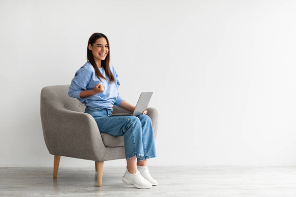 Vrolijke jonge vrouw zit in de fauteuil met laptop, het maken van YES gebaar, het krijgen van goed nieuws online, het bereiken van succes - Foto, afbeelding