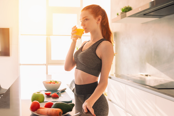 Athletic young red haired woman in the kitchen drinks a glass of fruit centrifuged juice - Zdjęcie, obraz