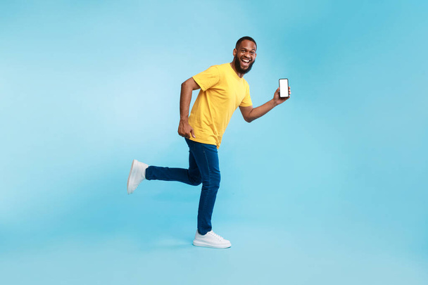 Young Afro guy with blank smartphone running on blue background, demonstrating mockup for mobile app, website - Photo, Image