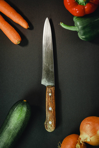 A vertical shot of a knife surrounded by peppers, carrots, zucchini, and onions - Foto, Bild