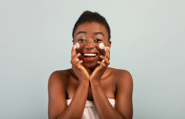 Denní rutina krásy. Šťastná africká americká dáma nanášení krém na tváře a úsměv na kameru přes šedé pozadí - Fotografie, Obrázek