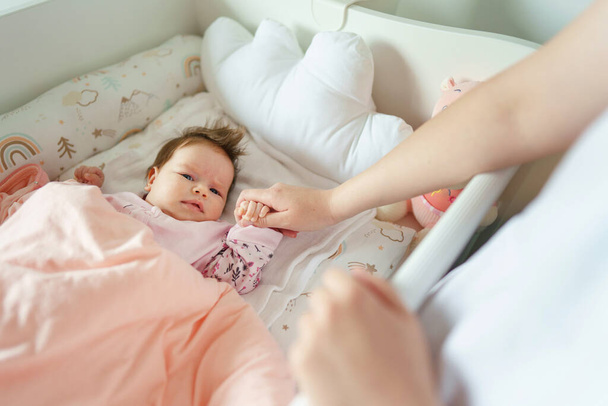 Top view on small Caucasian baby lying in cradle bed at home while her mother unknown woman is holding her hand putting her down to sleep motherhood care concept copy space - 写真・画像
