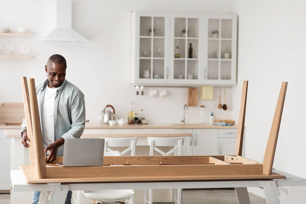 Millennial αφροαμερικανός πολυτεχνίτης συναρμολόγηση ξύλινο τραπέζι και κοιτάζοντας σε απευθείας σύνδεση οδηγίες στο laptop, αντίγραφο χώρου - Φωτογραφία, εικόνα