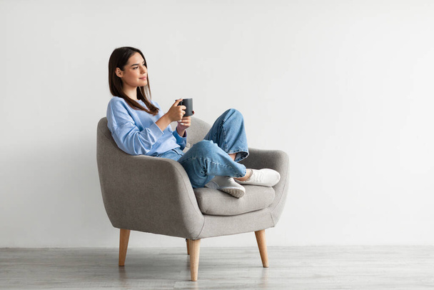 Positiivinen nuori nainen istuu nojatuolissa ristissä jalat, lämpeneminen kädet kupin kuumaa kahvia, nauttia lämmin juoma - Valokuva, kuva