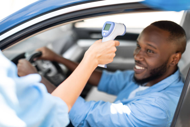 Ιατρικός εργαζόμενος που ελέγχει τη θερμοκρασία του σώματος του μαύρου άνδρα στο αυτοκίνητο - Φωτογραφία, εικόνα