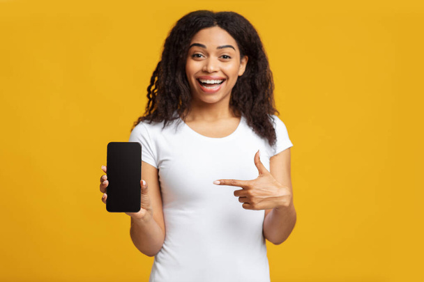 Positive black lady showing smartphone, pointing at empty blank screen, recommending mobile app, yellow background - Фото, изображение
